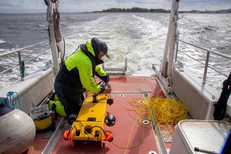 Bildet er tatt på dekket av en båt. Vi ser bakover på skummende hav i båtens kjølvann. På dekk står en mann i gul og blå kjeledress på huk mens han jobber med en gul, liten undervannsfarkost.