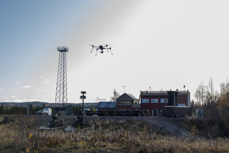 A drone hovering. I the backround two armoured vehicles, some houses, a crowd and a tall antenna. Autumn landscape in Norway.