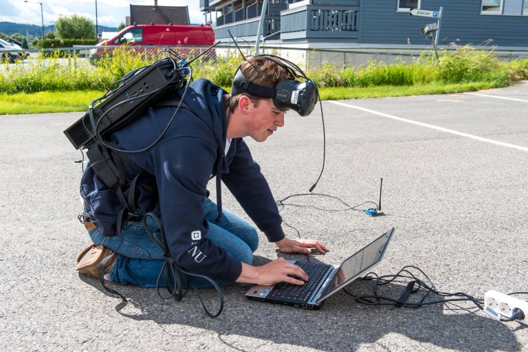 sommerstudent sitter ute med en laptop på bakken og utstyr i en sekk og på hodet